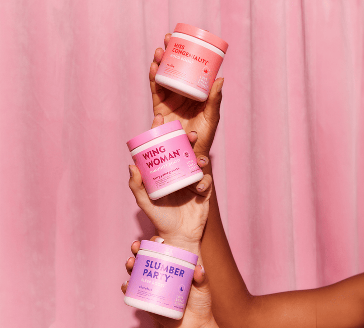 Three containers of wellness supplements in different flavors held up against a pink backdrop. The containers are labeled 'WING WOMAN', 'SLUMBER PARTY', and 'MISS CONGENIALITY'.