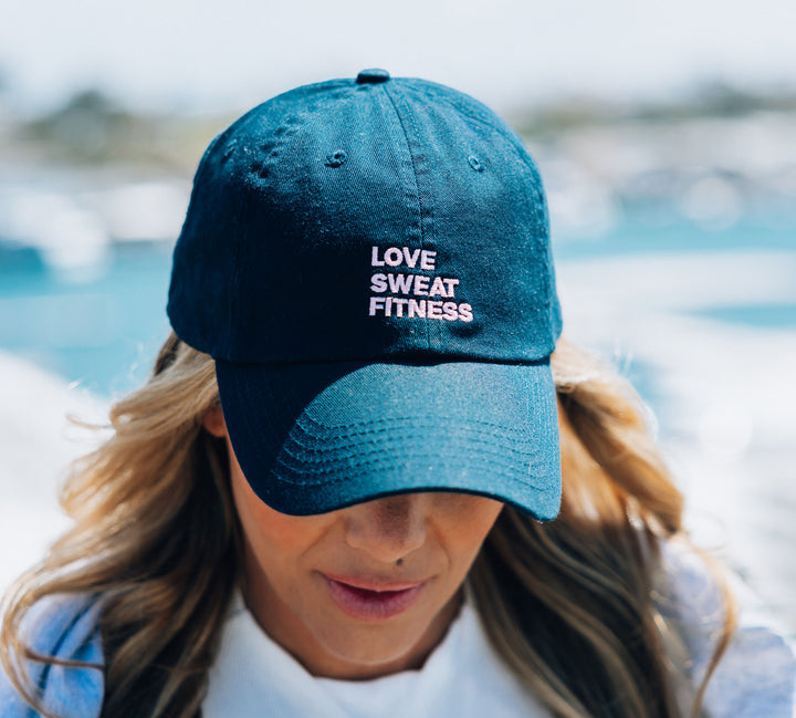 A person wearing a navy blue baseball cap with the embroidered text 'LOVE SWEAT FITNESS' on the front.